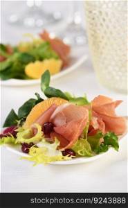 Portioned appetizer of prosciutto ham served on a of mixed with orange and pomegranate.