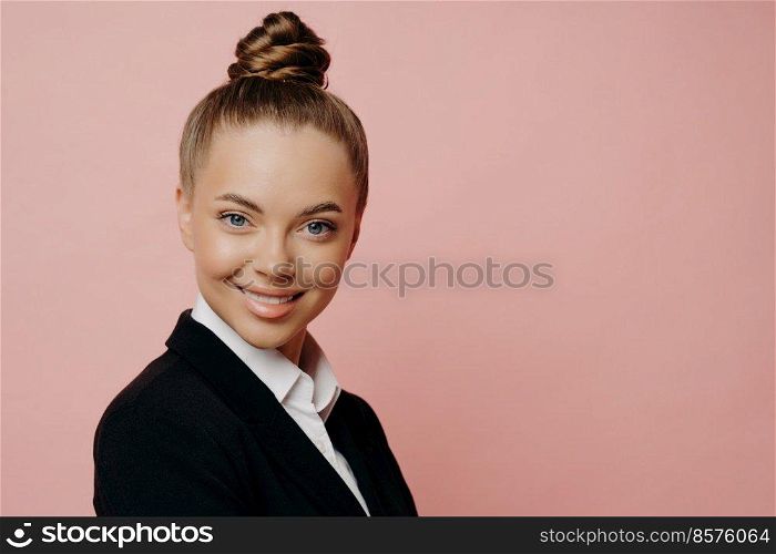 Portirat of cheerful happy female office employee in dark suit with hair in bun being notified about her promotion, with lovely smile standing sideways looking at camera, isolated over pink background. Half length shot portrait of happy pretty business woman