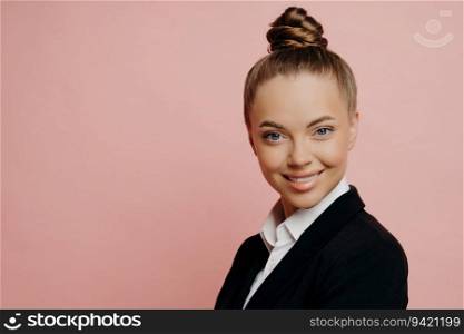 Portirat of cheerful happy female office employee in dark suit with hair in bun being notified about her promotion, with lovely smile standing sideways looking at camera, isolated over pink background. Half length shot portrait of happy pretty business woman
