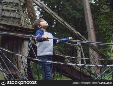 Portrait active kid climbing on rope frame at treehouse.Young boy standing alone and looking up at high forest tree near the park, Child playing outdoor playground alone in Autumn, .