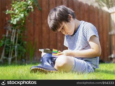 Portrait active little boy playing with construction plastic toy blocks in the garden, Child sitting on green grass looking deep in though while holding his toys, Kid playing outside in sunny morning, Toddler development concept