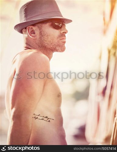 Portrait af a sexy shirtless sailor wearing hat and sunglasses enjoying sunset, man traveling along sea on luxury sailboat, happy summer vacation