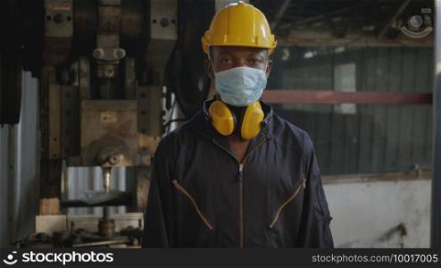 Portrait American industrial black young worker man smiling with yellow helmet and ear protection in front machine, Happy engineer wearing face mask standing at work in industry factory.