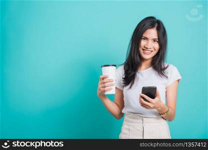 Portrait Asian beautiful happy young woman standing smile, using mobile phone her holding coffee paper cup, looking to camera on blue background, with copyspace