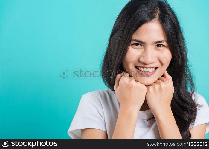 Portrait Asian beautiful happy young woman wear t-shirt glad keeps both hands under chin smiles pleasantly, on blue background, with copy space