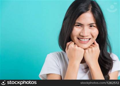 Portrait Asian beautiful happy young woman wear t-shirt glad keeps both hands under chin smiles pleasantly, on blue background, with copy space