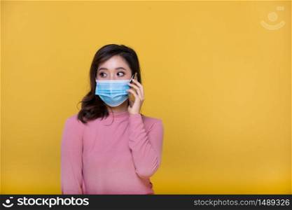 Portrait Asian beautiful happy young woman wearing face mask or protective mask against coronavirus crisis or COVID-19 outbreak and she is using mobile phone or smartphone on yellow background