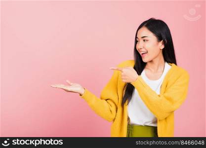 Portrait Asian beautiful young woman smiling standing pointing finger out on pink background, Thai happy face excited female point into empty looking to side away with copy space for text