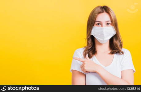 Portrait Asian beautiful young woman wearing face mask protects filter dust pm2.5 anti-pollution, anti-smog, air pollution and COVID virus her pointing out side on yellow background, with copy space