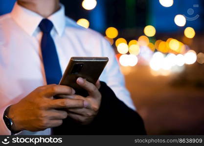 Portrait Asian businessman typing an sms message via smartphone after work near office at night city street, bokeh lights, young man walking and chatting with friends at social networks outdoor