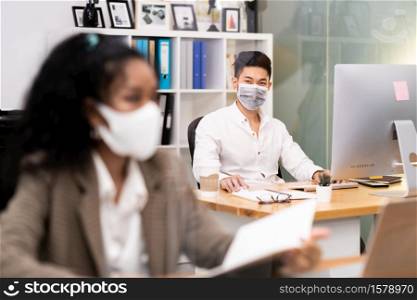 Portrait asian Office employee with protective face mask working in new normal office with social distance practice to american african colleague in foreground prevent coronavirus COVID-19 spreading.