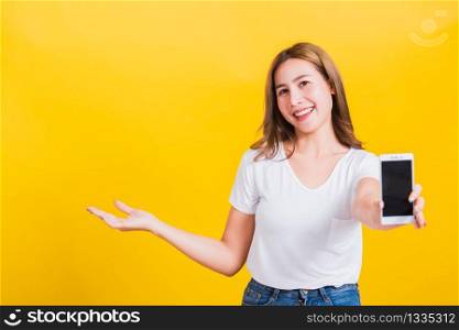 Portrait Asian Thai beautiful happy young lifestyle woman stands holding mobile phone and show something on side hand looking to camera, studio shot isolated on yellow background, with copy space