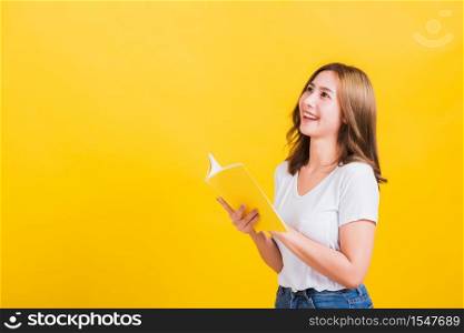 Portrait Asian Thai beautiful happy young lifestyle woman stands holding yellow book or diary she show thumb up finger and looking up away, studio shot isolated on yellow background, with copy space