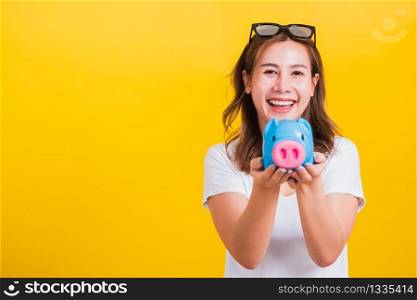 Portrait Asian Thai beautiful happy young woman smiling hold piggy bank with a lots money and looking to camera, studio shot isolated on yellow background, with copy space