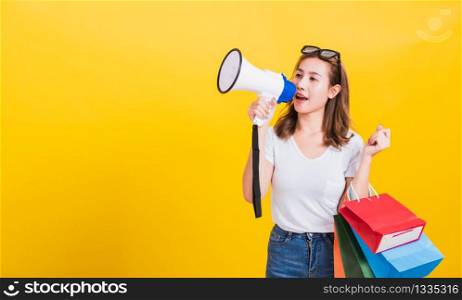 Portrait Asian Thai beautiful happy young woman smiling hold shopping bags multi-color and shouting in megaphone her looking to camera, studio shot isolated on yellow background, with copy space