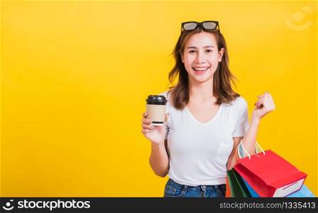 Portrait Asian Thai beautiful happy young woman smiling hold shopping bags multi-color and take away coffee cup her looking to camera, studio shot isolated on yellow background, with copy space