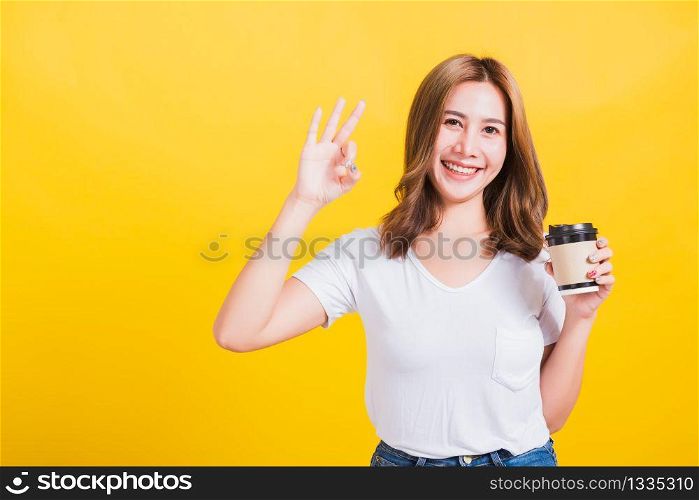 Portrait Asian Thai beautiful happy young woman standing holds take away cup coffee paper and confident show OK sign with fingers excellent, studio shot isolated on yellow background, with copy space