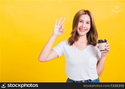 Portrait Asian Thai beautiful happy young woman standing holds take away cup coffee paper and confident show OK sign with fingers excellent, studio shot isolated on yellow background, with copy space