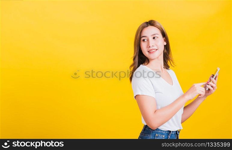 Portrait Asian Thai beautiful happy young woman standing smile, holding mobile phone her looking back to space, studio shot isolated on yellow background, with copyspace