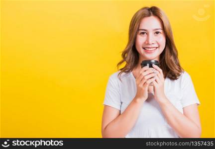 Portrait Asian Thai beautiful happy young woman wear white t-shirt standing smiling holding take away coffee paper cup, studio shot isolated on yellow background, with copy space