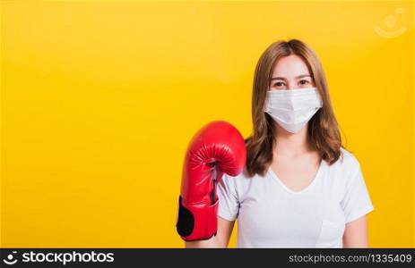 Portrait Asian Thai beautiful happy young woman wearing mask protection and boxing gloves her get ready for Coronavirus fighting against, studio shot isolated on yellow background, with copy space