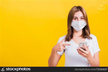 Portrait Asian Thai beautiful happy young woman wearing mask protection, pointing finger on smartphone health care, studio shot isolated on yellow background, with copy space