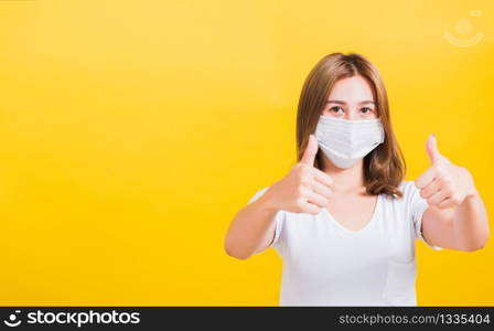 Portrait Asian Thai beautiful happy young woman wearing mask protection, show thumb finger good stopping virus outbreak control health care, studio shot isolated on yellow background, with copy space