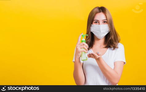 Portrait Asian Thai beautiful young woman wear mask protection clean corona virus on hand with alcohol gel or antibacterial soap sanitizer, studio shot isolated on yellow background, with copy space