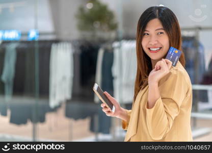 Portrait Asian woman using credit card with smart mobile phone for online shopping in department store over the clothes shop store background, technology money wallet and online payment concept