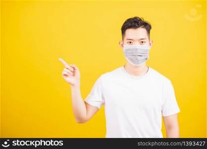 Portrait Asian young man wearing face mask protects filter dust pm2.5 anti-pollution, anti-smog, air pollution and COVID virus her pointing out side on yellow background, with copy space