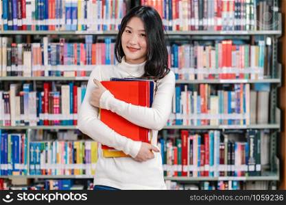 Portrait Asian young Student in casual suit standing and holding the book at book shelf in library of university or colleage with various book background, Back to school concept