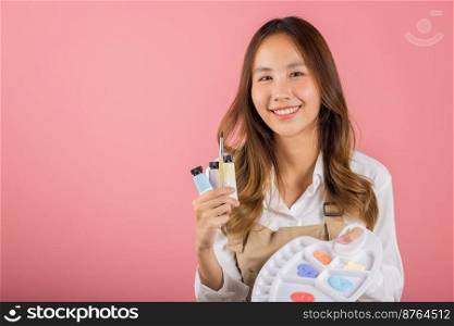 Portrait Asian young woman artist holding brush and paint palette, Happy female painting using paintbrush and palette with colors, studio shot isolated on pink background, Art design workshop