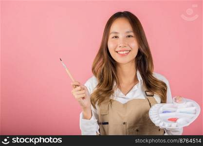 Portrait Asian young woman artist holding brush and paint palette, Happy female painting using paintbrush and palette with colors, studio shot isolated on pink background, Art design workshop