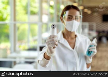 Portrait attractive asian waitress wear face mask and face shield hold thermometer and alcohol gel sanitizer for cleaning and hygiene with indoor restaurant background. New normal restaurant concept.