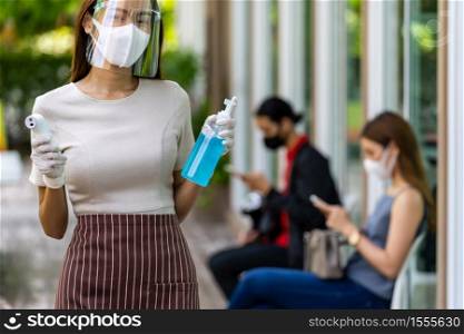 Portrait attractive asian waitress wear face mask and shield hold thermometer and alcohol gel sanitizer with background of social distancing customer waiting in queue. New normal restaurant concept.