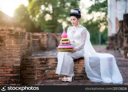 Portrait Beautiful asian woman with Thai white traditional dress costume holding Krathong and sitting in front of Pagoda temple at the ancient city Thailand,Loy Krathong Festival,Transgender model