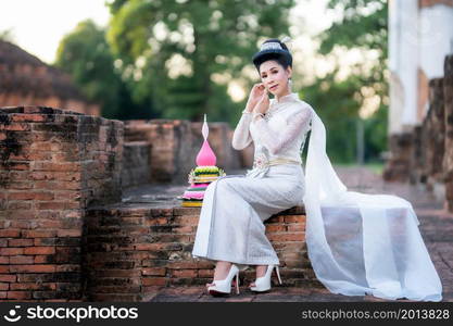 Portrait Beautiful asian woman with Thai white traditional dress costume holding Krathong and sitting in front of Pagoda temple at the ancient city Thailand,Loy Krathong Festival,Transgender model