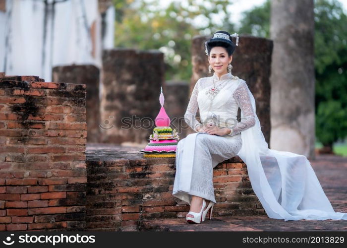 Portrait Beautiful asian woman with Thai white traditional dress costume holding Krathong and sitting in front of Pagoda temple at the ancient city Thailand,Loy Krathong Festival,Transgender model