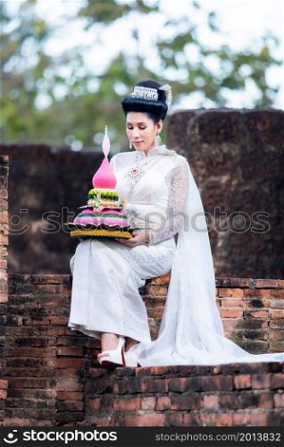 Portrait Beautiful asian woman with Thai white traditional dress costume holding Krathong and sitting in front of Pagoda temple at the ancient city Thailand,Loy Krathong Festival,Transgender model