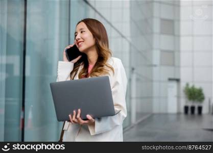 Portrait beautiful business woman smiling holding computer discussing issues on smart mobile phone in city, Asian businesswoman working on laptop and talking cell phone at front building near office