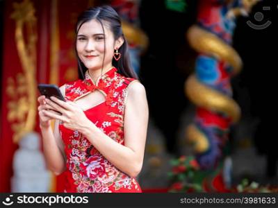 Portrait beautiful smiles Asian young woman wearing red cheongsam dress traditional decoration and writing message on smartphone for Chinese New Year Festival at Chinese shrine in Thailand