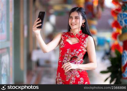 Portrait beautiful smiles Asian young woman wearing red traditional Chinese cheongsam decoration taking a selfie with smartphone for Chinese New Year Festival at Chinese shrine