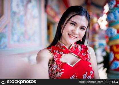 Portrait beautiful smiles Asian young woman wearing red traditional Chinese cheongsam decoration taking a selfie with smartphone for Chinese New Year Festival at Chinese shrine