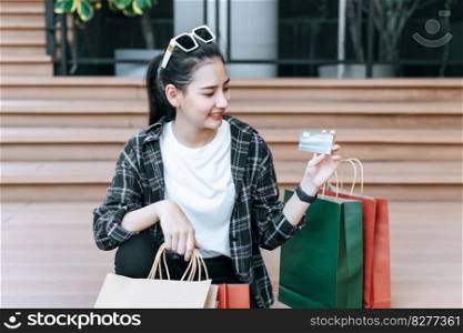 Portrait beautiful woman place eyeglasses on head holding shopping bags in hand sit on stairs of mall, she smile and show credit cards in hand,  Lifestyle and shopping concept