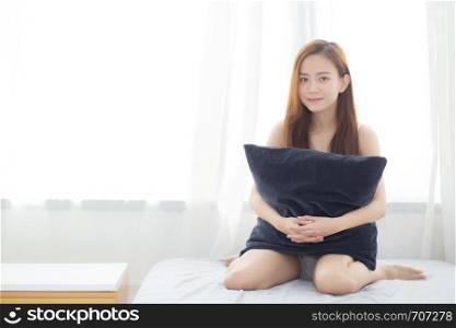 Portrait beautiful young asian woman sitting and smile hug pillow with the window at bedroom while wake up with sunrise at morning, lifestyle and relax concept.