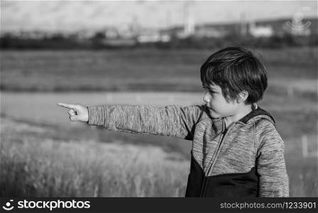 Portrait black and white photo of happy little boy pointing finger up , Active kid having fun playing in farm field with bright light sunny day. , Child playing outdoor with blurry natural background.