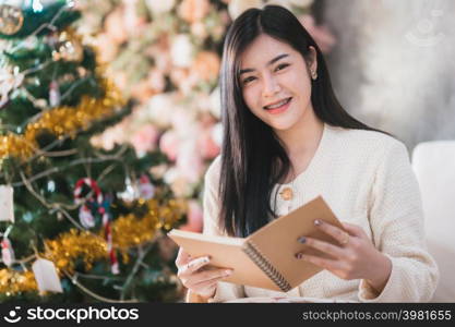 Portrait Cute beautiful positive smile young asian woman holding of read a book at home in the living room indoors Decoration During Christmas x-mas and New Year holidays.