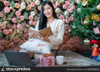 Portrait Cute beautiful positive smile young asian woman holding of read a book and working with laptop at home in the living room indoors Decoration During Christmas x-mas and New Year holidays.