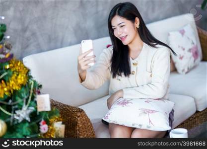 Portrait Cute beautiful positive smile young asian woman using smartphone video call or Selfie at home in the living room indoors Decoration During Christmas x-mas and New Year holidays.