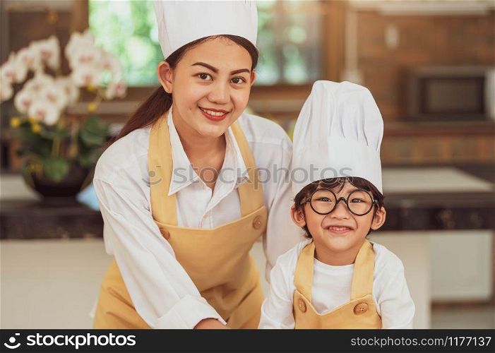 Portrait cute little Asian boy chef with eyeglasses and his mother looking to camera in home cooking kitchen happily and funny. People lifestyles and Family. Homemade food ingredient making concept